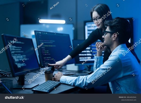 A manager helping a worker by pointing someone out to them on a monitor.